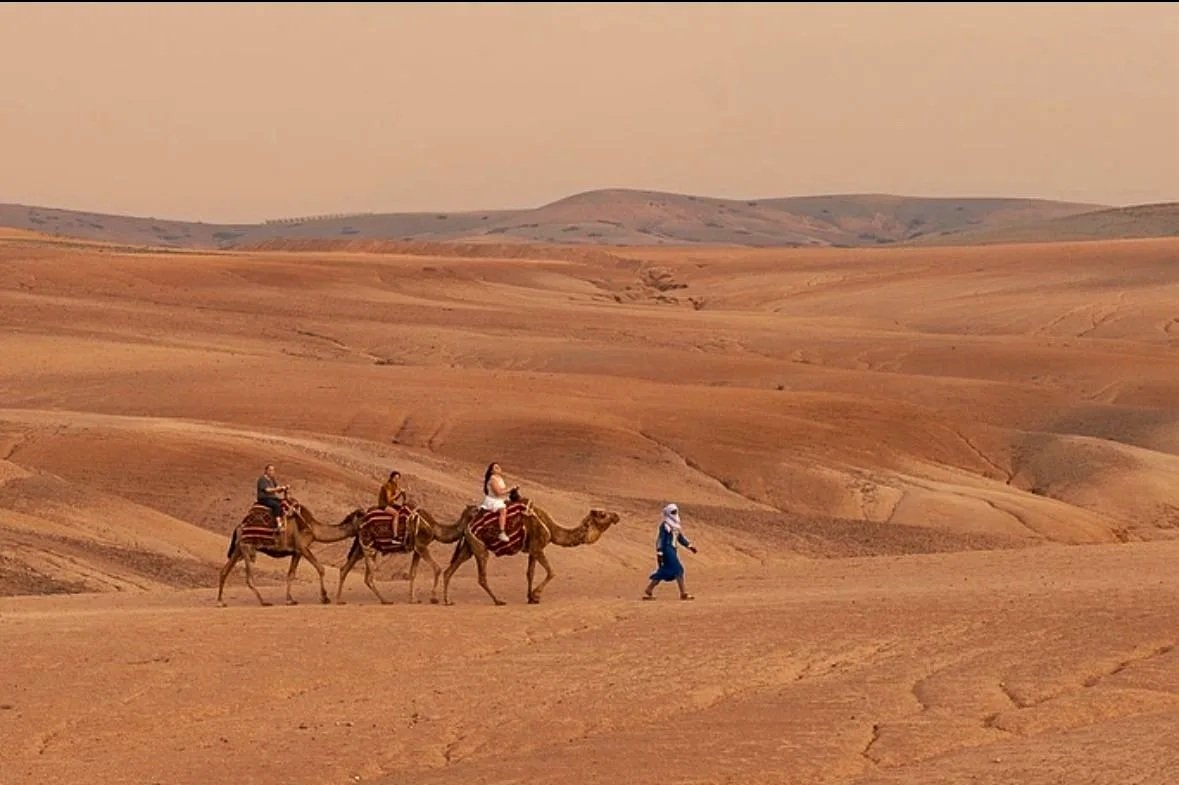 Group of three people riding camels across a desert landscape with rolling sand dunes, accompanied by a person walking on foot.