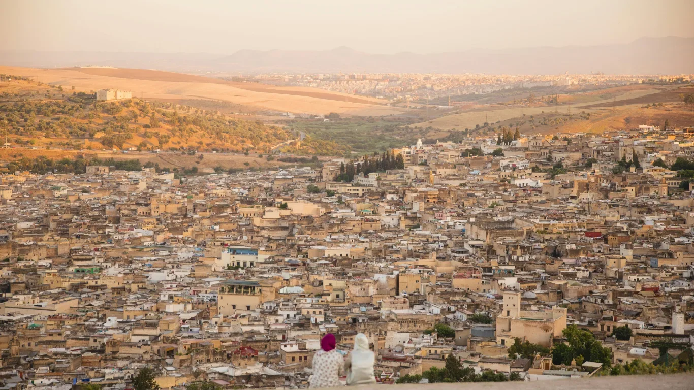 Two women sit on a hill overlooking a densely populated city with many beige buildings, surrounded by mountains and rolling hills in the background.