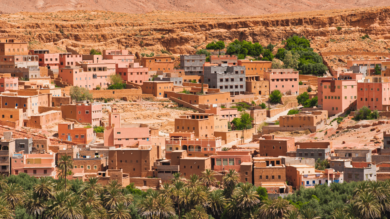 A desert hillside landscape with numerous colorful buildings, palm trees in the foreground, and rugged rock formations in the background.