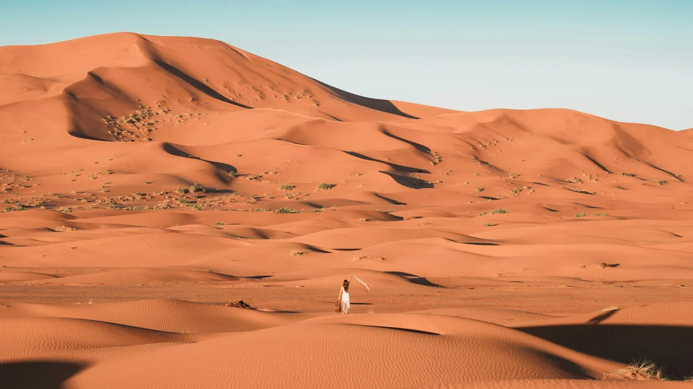 A woman in a white dress walking across a desert with orange sand dunes and sparse vegetation, under a clear blue sky.