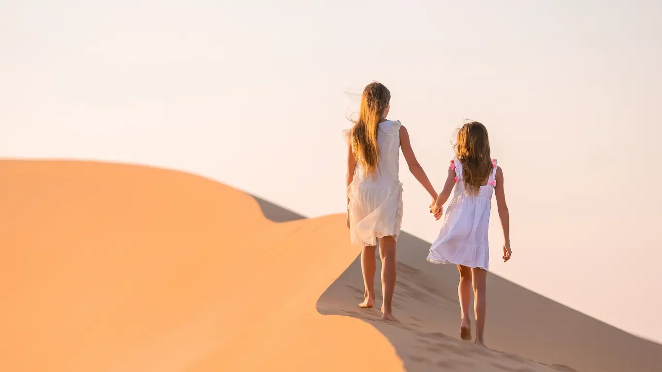 Two girls in white dresses holding hands and walking on a sand dune in a desert at sunset.