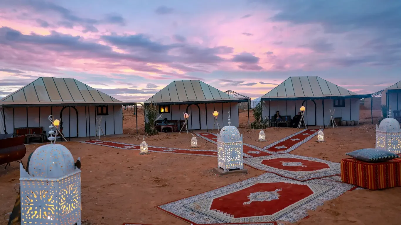 A desert campsite with four white tents, decorated with patterned rugs on the sand, lanterns, and lantern-shaped lamps, under a colorful sunset sky.