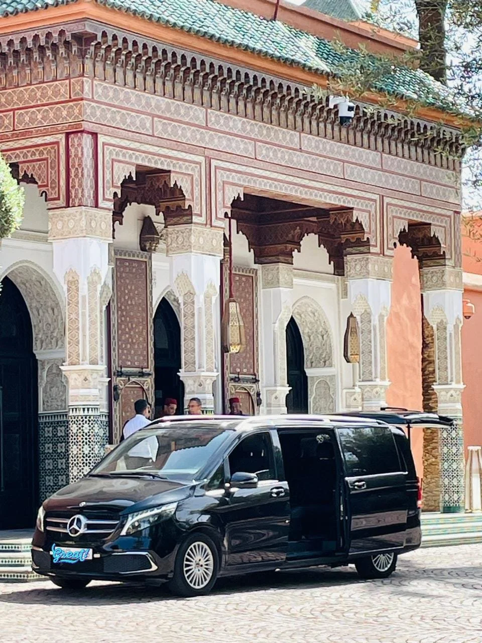A black Mercedes-Benz van parked in front of a building with ornate, colorful Islamic architecture, including arches, intricate tile work, and hanging lanterns. Several people are seen near the entrance.