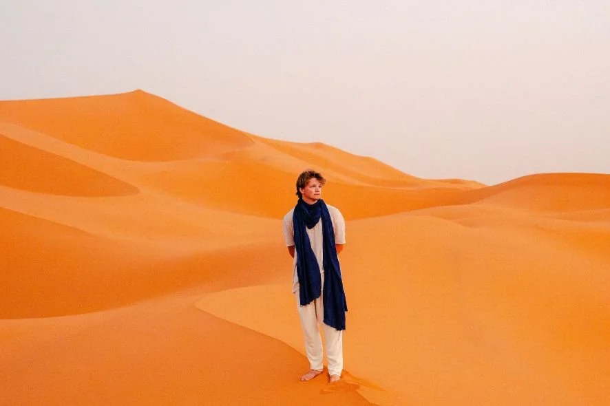 Person standing in a desert among sand dunes, wearing light-colored clothing and a dark blue scarf.