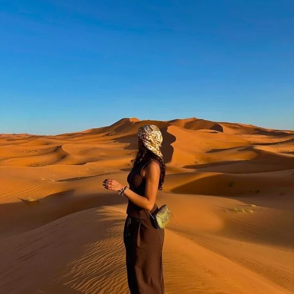 Woman walking in a desert with sand dunes and clear blue sky.