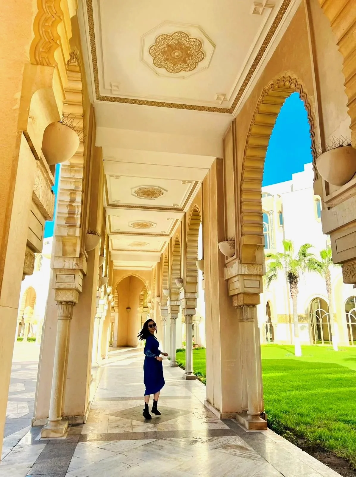 A woman in a blue dress and black boots walking through an ornate, sunlit corridor with arches, columns, and intricate ceiling designs, surrounded by a well-maintained grassy area and palm trees.