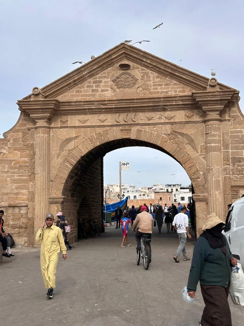 People walking and biking through an old stone archway, with buildings in the background, seagulls flying overhead, and some boats docked nearby.