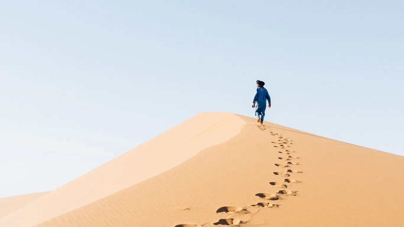 A person in blue clothing walking along the crest of a sand dune in a desert, leaving footprints behind.
