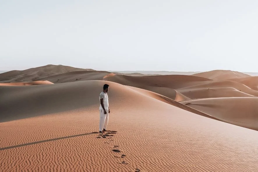 A man dressed in white standing in a desert with sand dunes, leaving footprints behind.