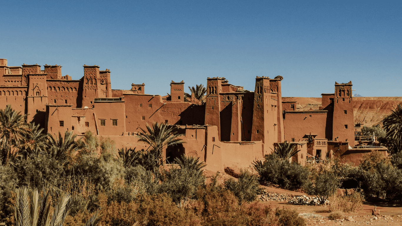 A desert fortress with high mud brick walls and towers, surrounded by palm trees under a clear blue sky.