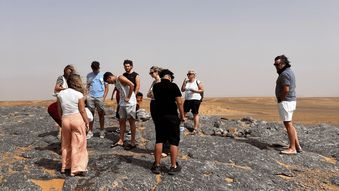 Group of tourists standing on rocky terrain in a desert, facing each other and listening as a guide explains, with a vast flat desert landscape in the background.