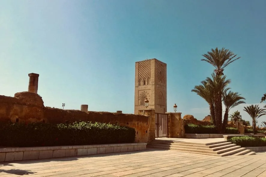 A historic Moroccan tower surrounded by palm trees, with a stone staircase, a fence, and lanterns, under a clear blue sky.