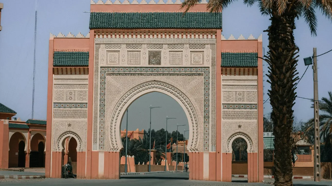 A large ornate archway with geometric and floral patterns, painted in pink and white, with green tiled roofing. There are smaller archways and palm trees around, and street lamps visible in the background.