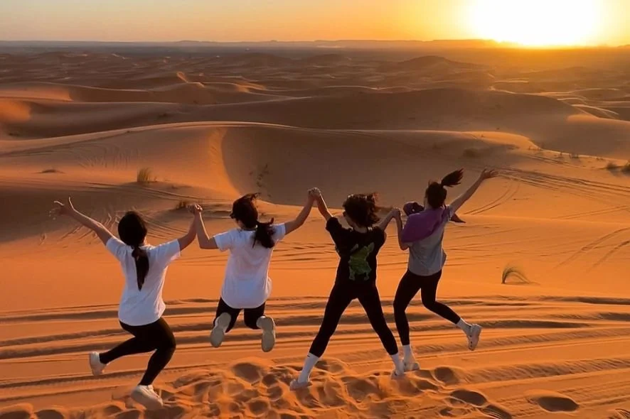 Four people holding hands and jumping on sand dunes at sunset in a desert.