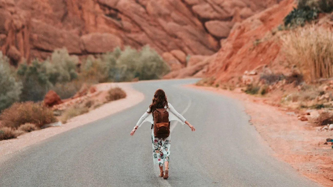 A woman walking on a deserted winding road in a desert landscape with red rocks and sparse vegetation.