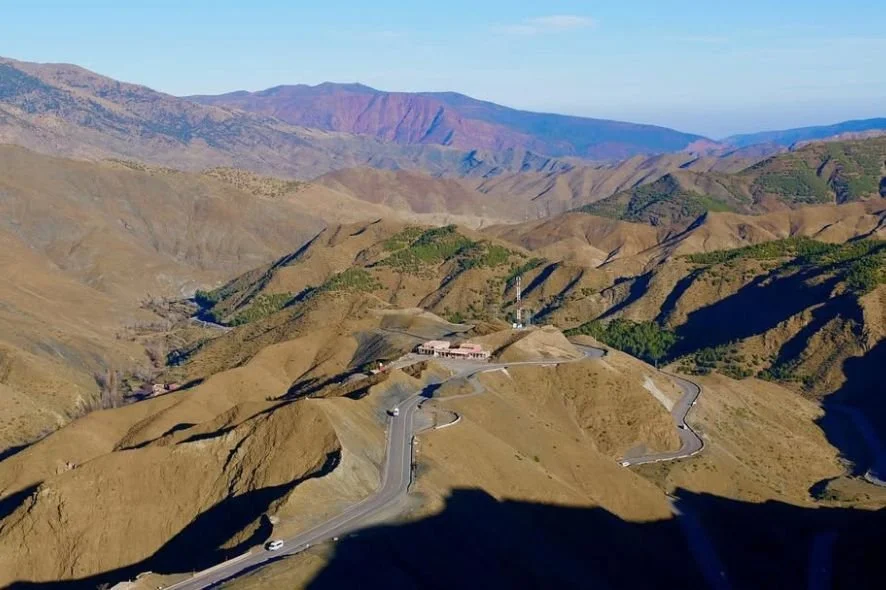 A winding mountain road with a vehicle traveling on it, surrounded by brown, barren hills and distant colorful mountains under a blue sky.
