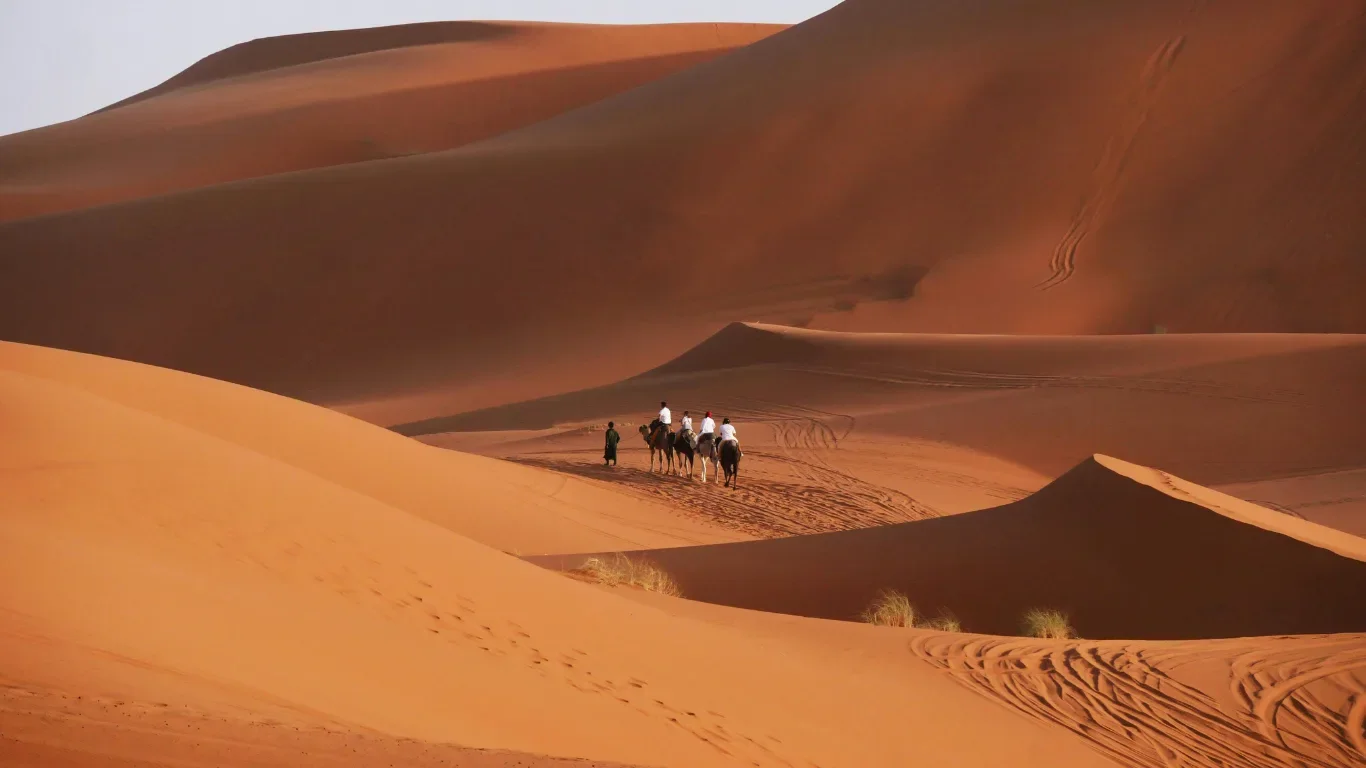 Four people riding camels through a vast desert with towering sand dunes, minimal vegetation, and parallel tracks in the sand.