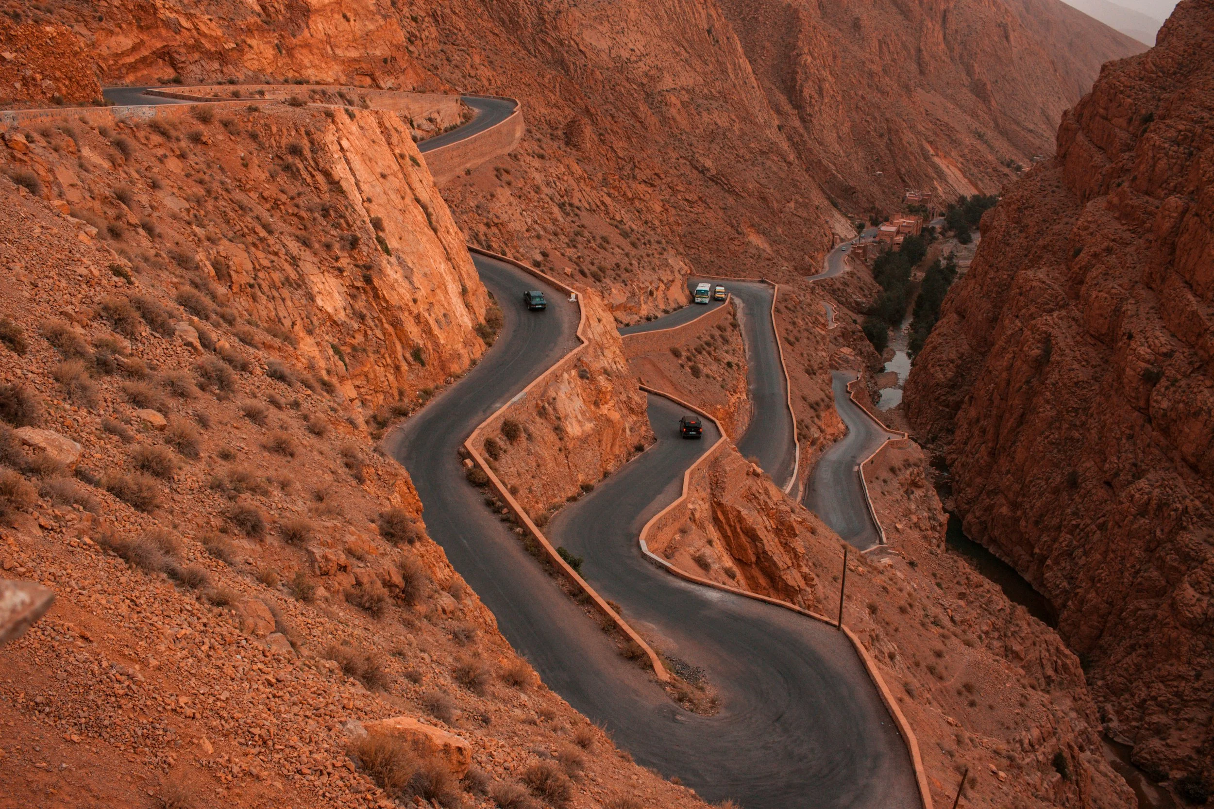 A winding road zigzags through rugged, reddish-orange desert canyon cliffs, with several vehicles traveling along it.