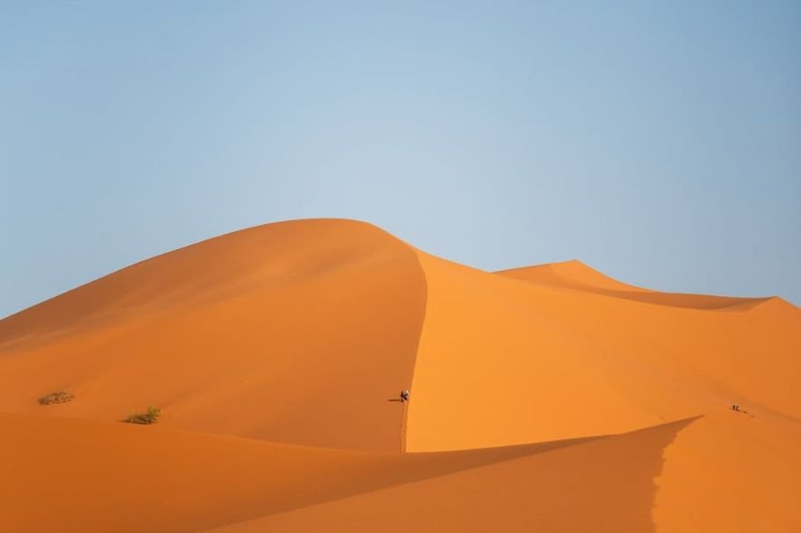 A vast desert landscape with large orange sand dunes against a clear blue sky, with a person riding a camel along the crest of a dune.