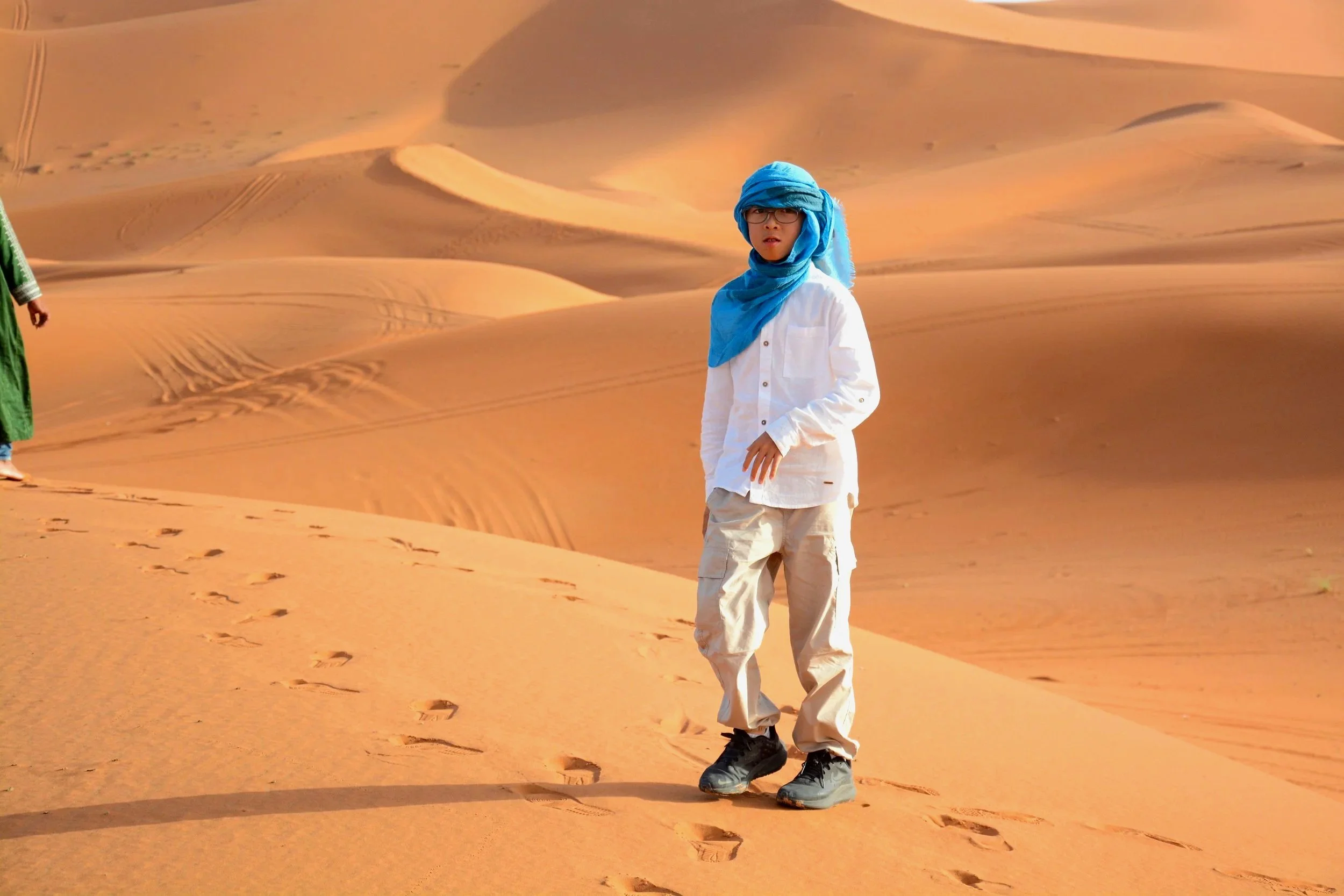 A person standing on sand dunes in a desert, dressed in beige cargo pants, a white shirt with sleeves rolled up, a blue headscarf, glasses, and black hiking shoes, with footprints visible in the sand.