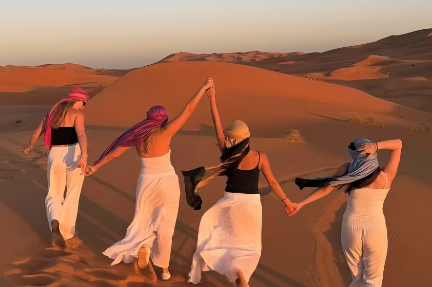 Four women hold hands and walk through a desert landscape with sand dunes at sunset.