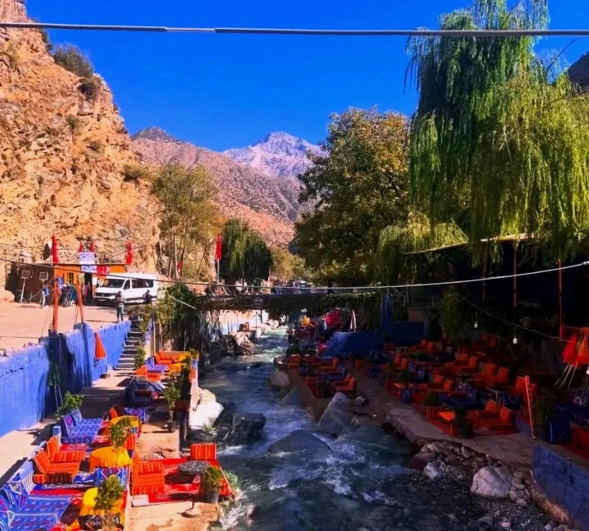 River running through a small town with colorful seating and tables on both sides, surrounded by trees and mountains under a clear blue sky.