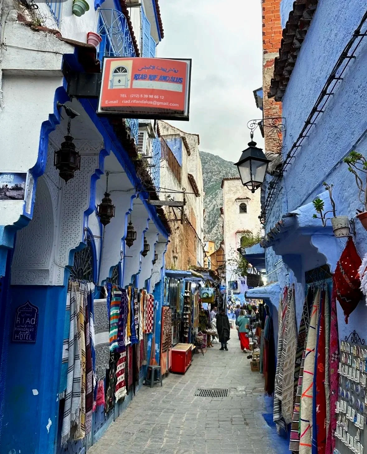 A street market in a blue-painted town, with colorful textiles on display on both sides, hanging from shop fronts. People are walking along the cobblestone street, and there are hanging lanterns and potted plants visible.