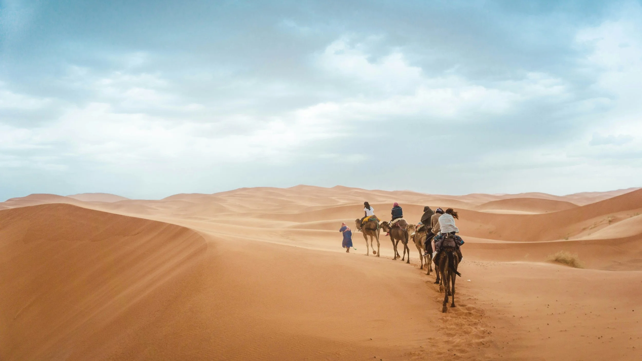 Group of people riding camels across sand dunes in a desert with cloudy sky.
