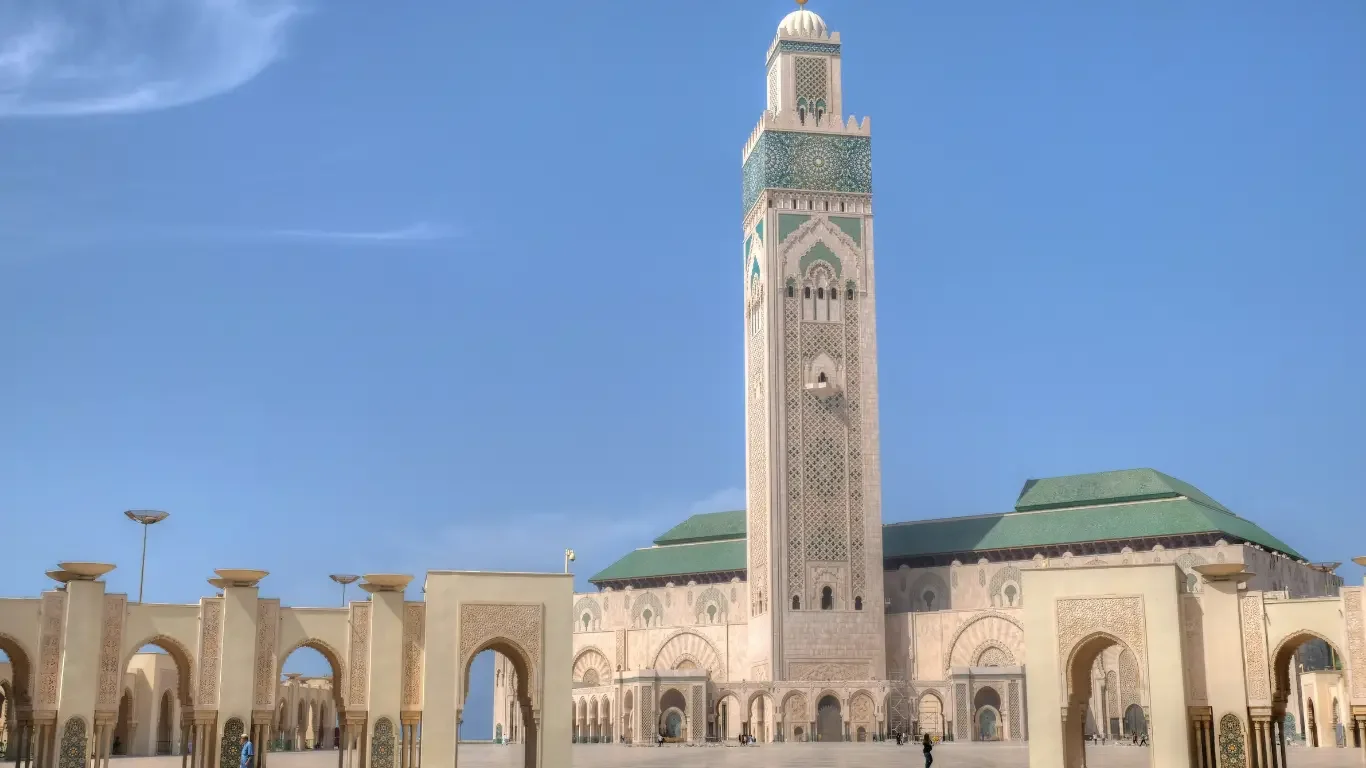 View of the Hassan II Mosque with its tall minaret against a blue sky and an open courtyard with arches in Casablanca, Morocco.