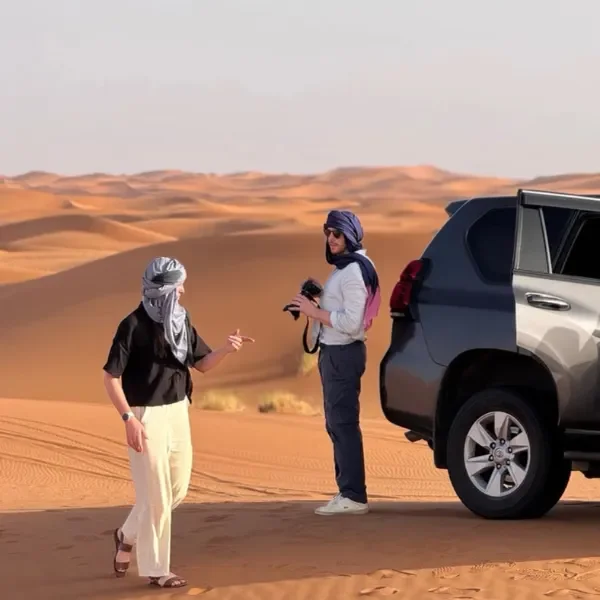 Two women having a conversation in a desert with sand dunes in the background. One woman is wearing a black top and beige pants, while the other woman is holding a camera and wearing a light-colored shirt and navy blue pants. A gray SUV is parked nearby.