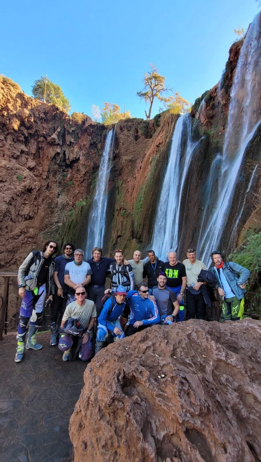 A group of people posing in front of a tall waterfall with multiple streams, on a sunny day with clear blue sky, surrounded by rocky terrain and sparse trees.