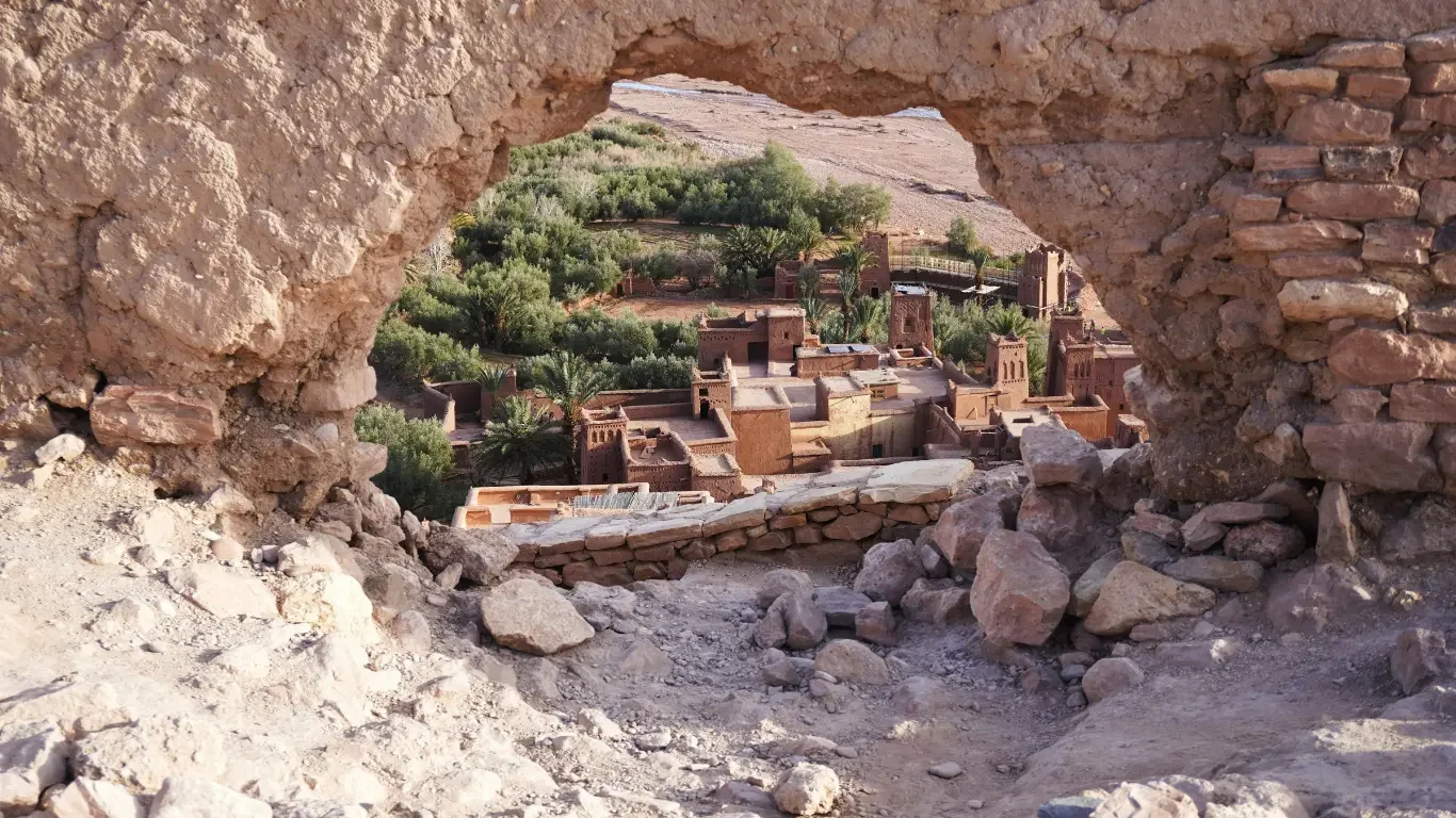 View through a stone window opening showing a traditional desert village with mud brick buildings and green trees.