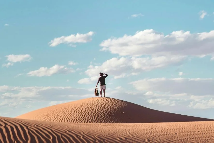 A person standing on a sand dune in a desert, holding a bag and looking at the sky with scattered clouds.