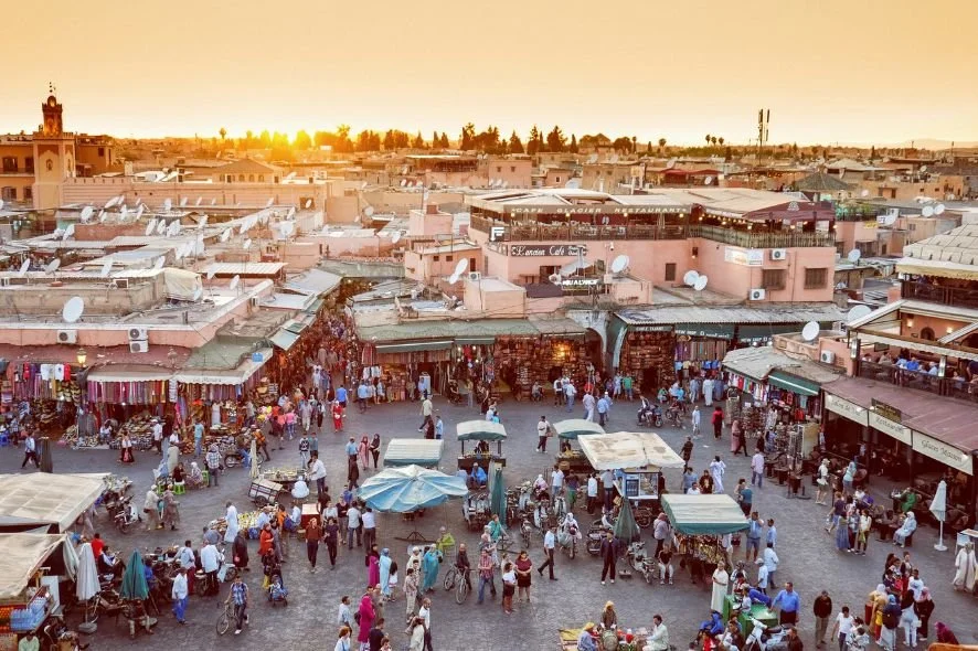 Busy outdoor marketplace in Morocco at sunset with shops, vendors, and shoppers.