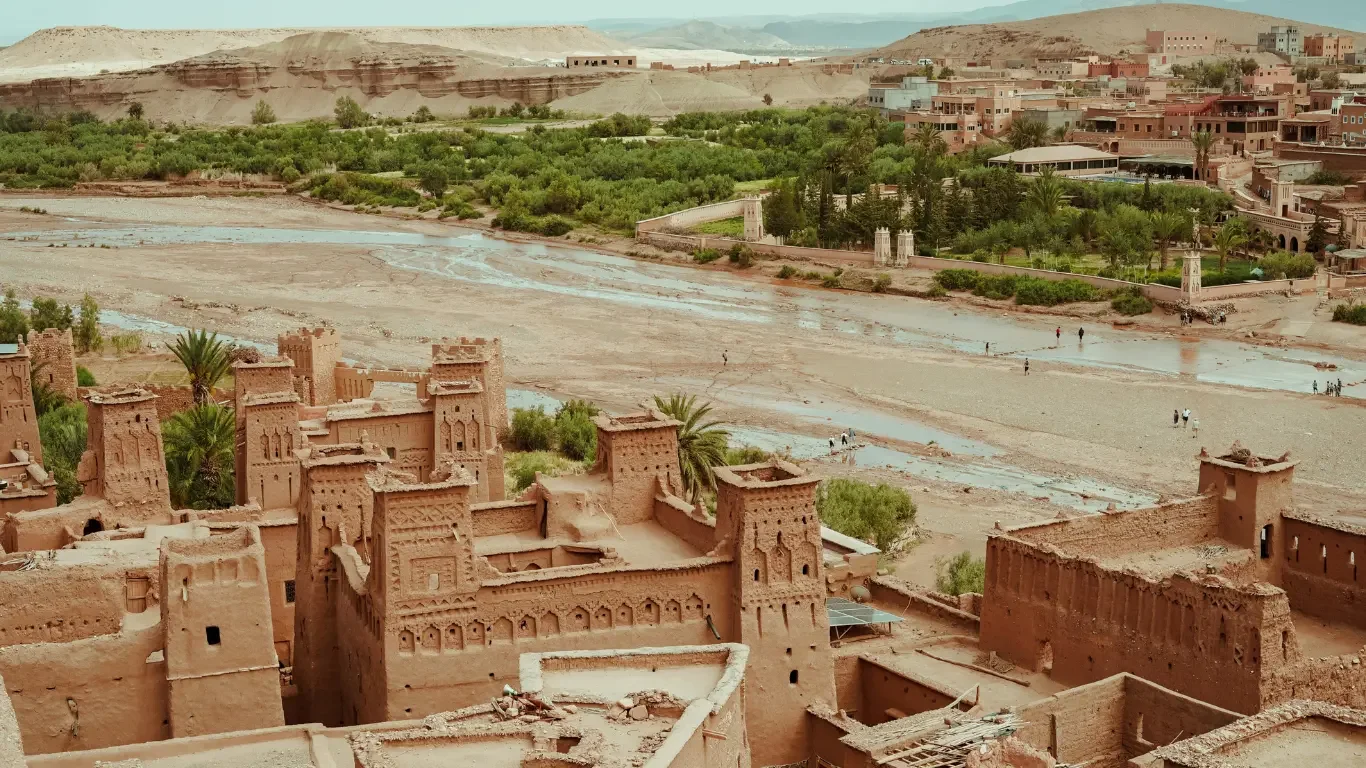 Ancient desert fortress with tall mud-brick walls and towers, surrounded by palm trees and a modern city with buildings, and a river in the background.