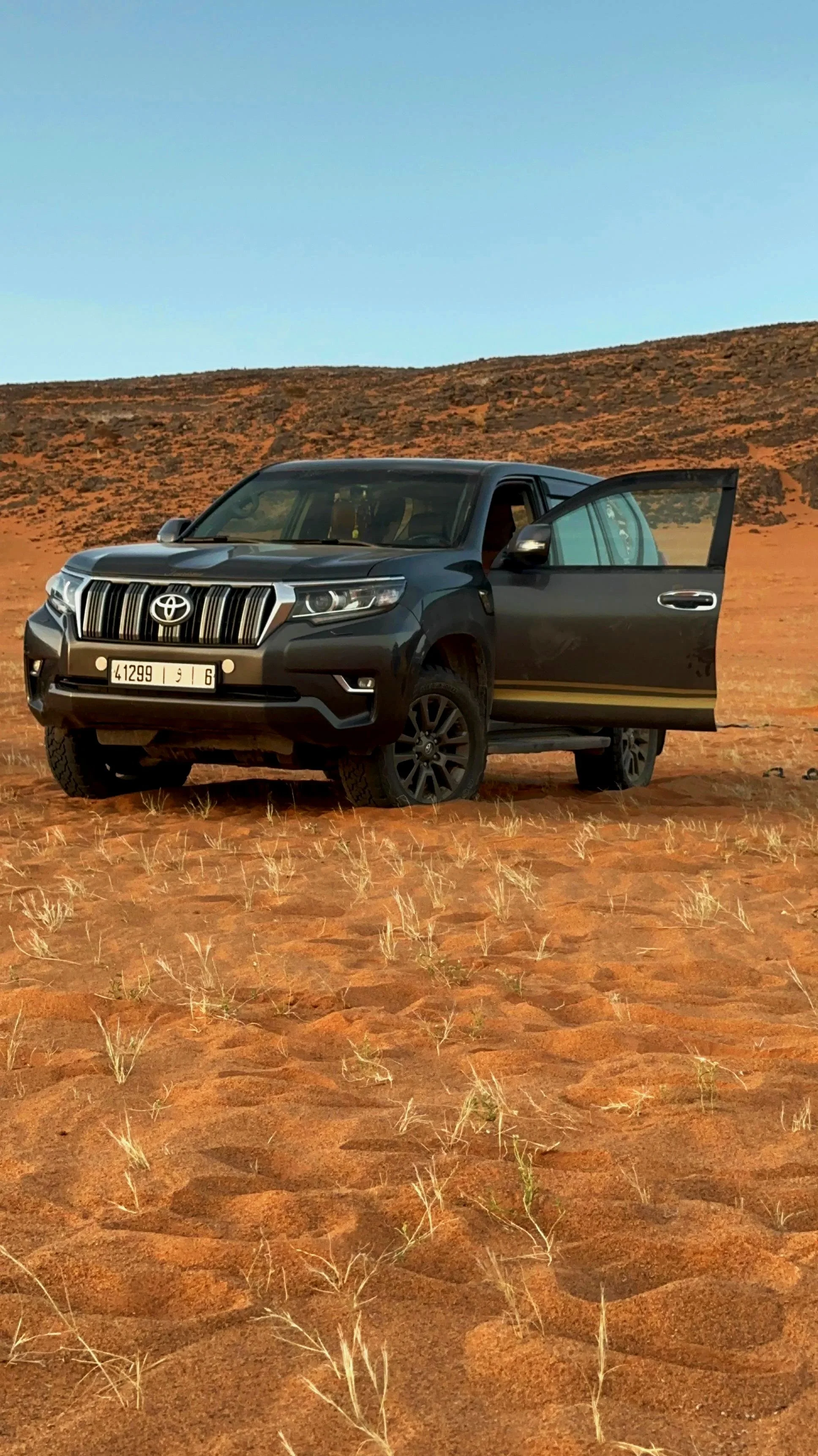 A black SUV parked on a dirt ground in a desert landscape with a large, old, damaged tent and a clear blue sky in the background.