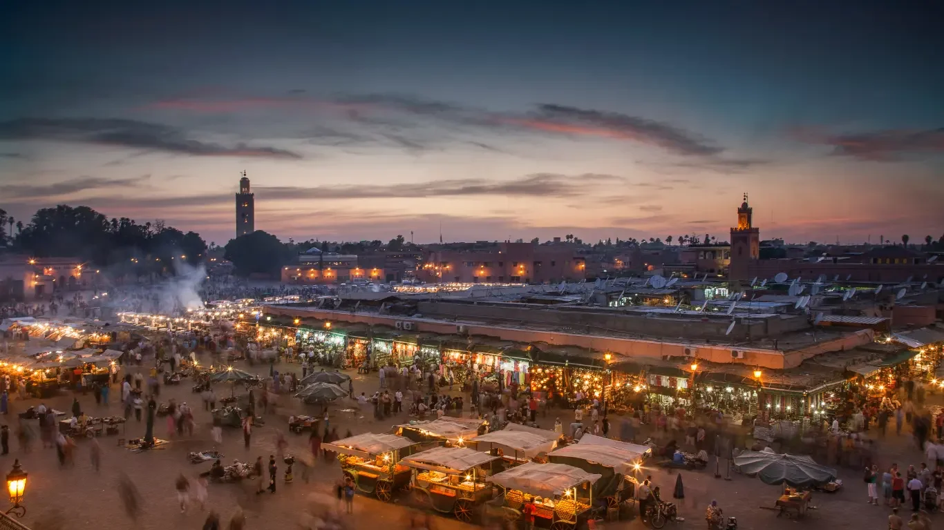 A lively marketplace at dusk with numerous small stalls, a crowd of people, and city buildings with tall towers in the background.
