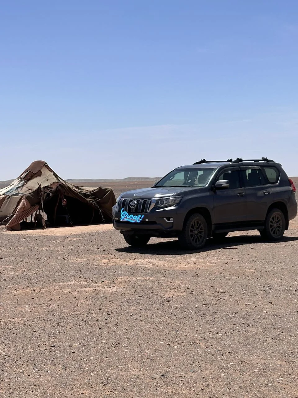 A black SUV parked on a dirt ground in a desert landscape with a large, old, damaged tent and a clear blue sky in the background.