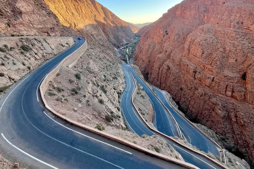 Winding road through a canyon with red rock formations on both sides, captured during sunset or sunrise.