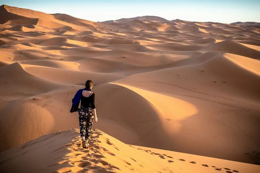 A woman walking across sand dunes in a desert with footprints behind her, under clear sky.