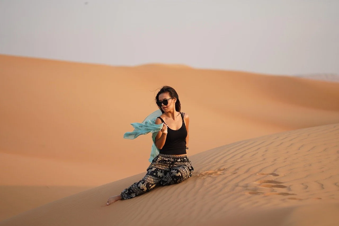 A woman wearing sunglasses and patterned pants kneeling on sand dunes in a desert, holding a light blue scarf.