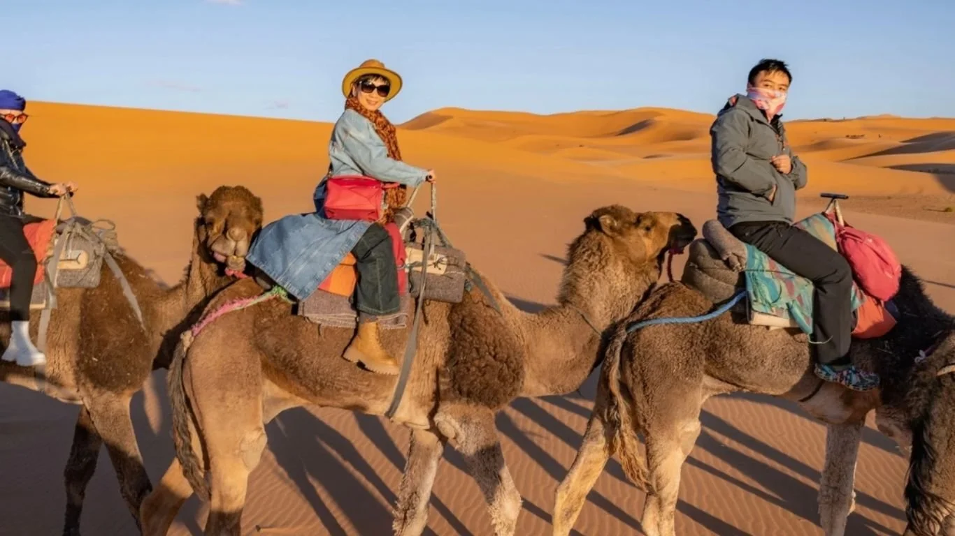 Two people riding camels in a desert with sand dunes in the background during sunset.