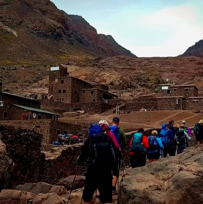 A group of hikers with backpacks trekking through a mountainous landscape with desert-like terrain and stone buildings in the background.