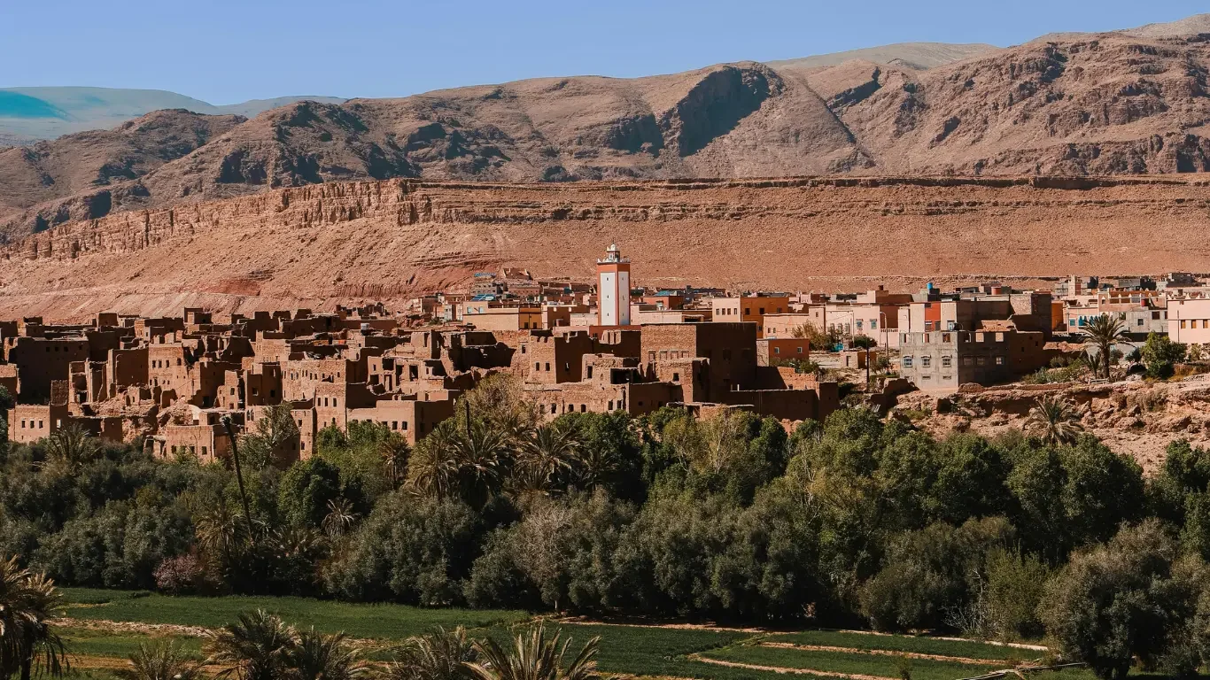 A town with traditional earthen buildings, set against a mountainous desert landscape, with lush greenery and palm trees in the foreground.