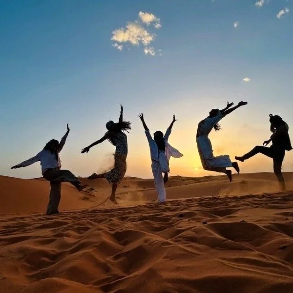 Five people silhouetted against a sunset sky, jumping and playing on sand dunes in a desert.
