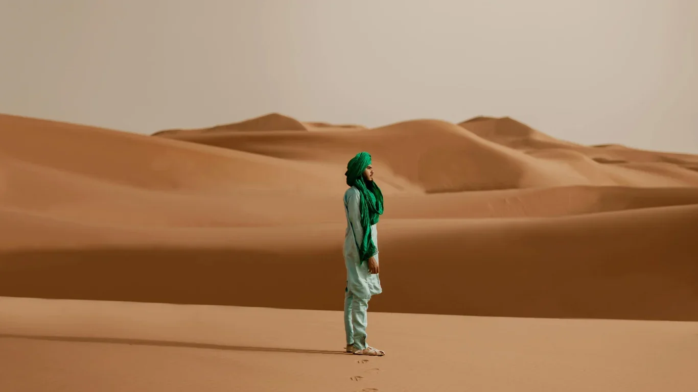 A woman dressed in traditional attire with a green headscarf and scarf, standing in a desert with sand dunes in the background.