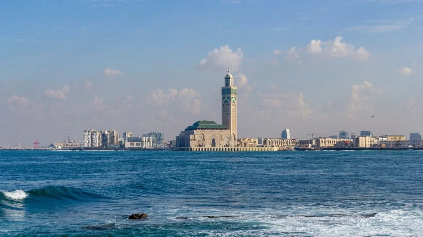 View of the city of Casablanca, Morocco, with the Hassan II Mosque prominently visible along the coastline, under a partly cloudy sky with the Atlantic Ocean in the foreground.
