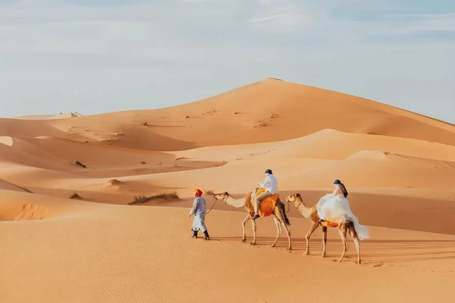 Two people riding camels in a desert with sand dunes and a cloudy sky.