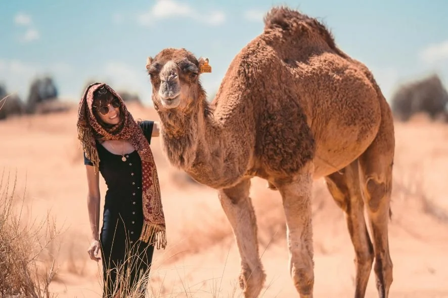 Woman with scarf and sunglasses standing next to a camel in a desert landscape.