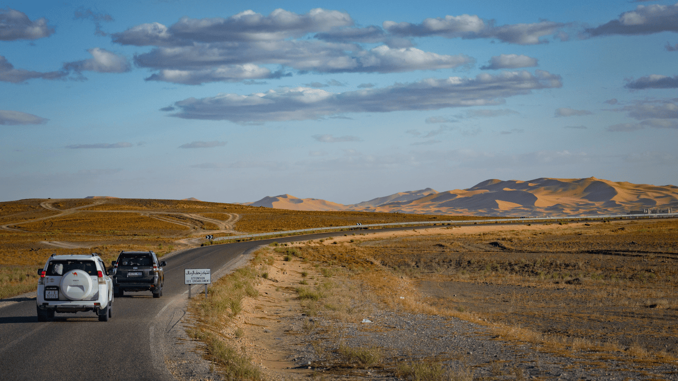 Cars driving on a winding road through a desert landscape with sand dunes in the background and a partly cloudy sky.